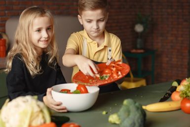 Positive little girl and boy mixing cutted ingredients for salad in big bowl while cooking at table with assorted fresh vegetables and fruits